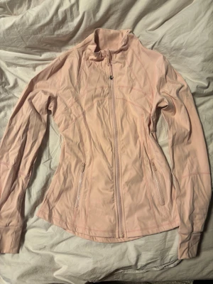 Ljusrosa vindjacka från Lululemon - Snygg ljusrosa vindjacka från Lululemon med figurnära passform. Jackan har hel dragkedja framtill, två sidofickor med dragkedja och högt krage. Materialet är mjukt och stretchigt syntet, perfekt för träning eller chill. Lululemon-logga på bröstet och ryggen.