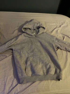 Grå hoodie från H&M med huva - Säljer en enkel och snygg grå hoodie från H&M i storlek 158/164. Tröjan har en klassisk huva, långärmade ärmar och en stor magficka framtill. Perfekt för chill dagar och passar till det mesta. Materialet är mjukt och skönt, troligen bomull och polyester.