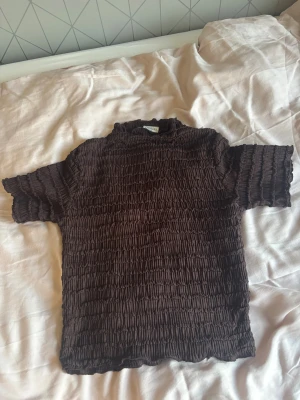 Brun smockad topp från Stories XS - Säljer en brun smockad topp från Stories i storlek XS. Toppen har korta ärmar, hög hals och är tillverkad i ett stretchigt material med smockad struktur som ger en cool och trendig look. Perfekt att styla med jeans eller kjol för en avslappnad vibe. Använd 1 gång