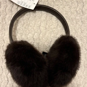 Bruna fluffiga öronmuffar i päls - Helt nya snygga och mjuka öronmuffar i mörkbrun färg. Perfekt för att hålla öronen varma under kalla dagar. Sitter bekvämt med ett böjbart band över huvudet. En stilren accessoar för vintern.