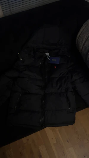 Svart pufferjacka från Polo Ralph Lauren - Svart pufferjacka från Polo Ralph Lauren med huva och dragkedja framtill. Jackan har två sidofickor med dragkedja och klassisk röd Polo-logga på bröstet. Perfekt för kalla dagar och riktigt snygg streetstil.