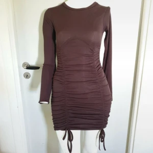 Brun ribbad omlottklänning med snörning - Snygg brun kortklänning med ribbad struktur och långa ärmar. Klänningen har omlott-effekt och snörning längs sidorna som ger en cool rynkad look. Passformen är tight och materialet känns mjukt mot huden. Perfekt för dig som gillar trendiga detaljer och vill sticka ut.