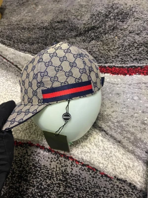 Gucci keps med GG-mönster och röd-blå rand - Snygg keps från Gucci i beige och marinblått med klassiskt GG-mönster över hela kepsen. På sidan finns en bred röd och marinblå rand. Justerbar rem baktill och insida i marinblå bomull. Perfekt för dig som vill ha en lyxig streetstyle-look.