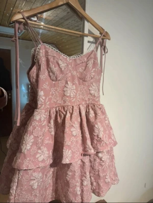 Rosa blommig kortklänning med volanger - Supersöt rosa kortklänning med vita blommor och volanger. Klänningen har tunna axelband med knyt. Har använt den en gång!💕