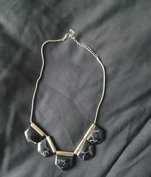 Svart och guldigt statement-halsband - Unikt halsband med fem svarta, geometriska hängen dekorerade med vita linjemönster. Kedjan är guldig och har justerbar längd. Perfekt för dig som vill sticka ut med en cool accessoar med modern känsla.