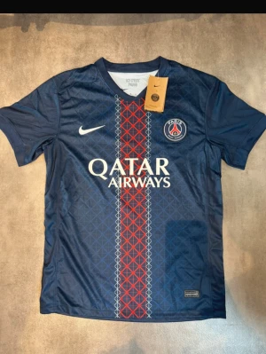 PSG matchtröja - Paris Saint-Germain matchtröja från Nike i marinblått med geometriskt mönster i rött och vitt längs mitten. Tröjan har korta ärmar, rund hals och är tillverkad i lätt Dri-Fit polyester för bästa komfort på planen. PSG-logga och Qatar Airways tryck på bröstet.