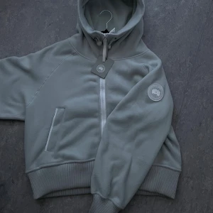 Ljusgrå fleecejacka från Canada Goose - Snygg ljusgrå fleecejacka från Canada Goose med huva, dragkedja och ribbade muddar. Jackan har två fickor framtill och en broderad logotyp på ärmen. Perfekt för kyliga dagar och har en clean, modern look. Öppen för förslag om priset, så hör gärna av er😁