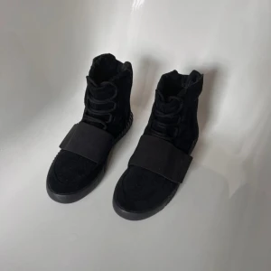 Adidas Yeezy Boost 750 Svarta - Säljer ett par svarta Adidas Yeezy Boost 750 med högt skaft, bred rem över foten och snörning. Skorna har en chunky, räfflad sula med boost-teknologi och är tillverkade i mocka för en lyxig känsla. Perfekt för dig som gillar streetwear och vill sticka ut.