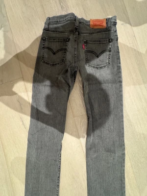 Grå Levi's 511 jeans straight fit - Snygga grå jeans från Levi's modell 511 med klassisk femficksdesign och raka ben. Jeansen har en mörkgrå tvätt med subtila slitningar och detaljer i kopparfärg. Perfekt för dig som gillar en clean och tidlös look. Storlek 10a. Andvänt fåtal gånger