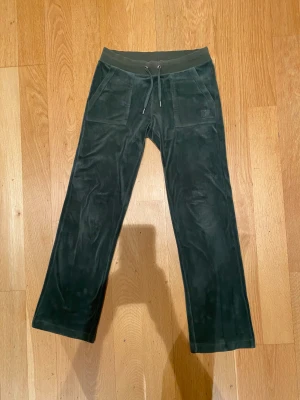 Mörkgröna mjukisbyxor Juicy Couture XS - Mörkgröna mjukisbyxor från Juicy Couture i supermjuk velour med bred resårmidja och snörning. Byxorna har raka ben, två stora fickor fram och bakfickor med knappdetalj. Perfekta för en chill och trendig look. Storlek XS men passar även S. Modell: Classic Velour Del Ray Pant Dark Moss.