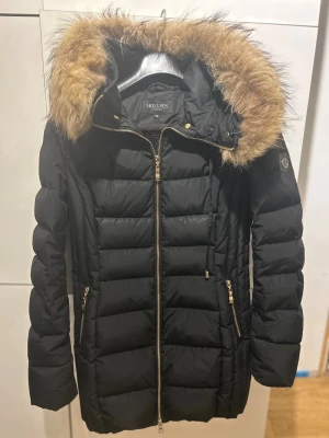 Svart dunjacka med äkta päls från Hollies - Snygg svart dunjacka från Hollies med guldig dragkedja och detaljer. Jackan har en fluffig huva med äkta päls, quiltad design och två fickor med dragkedja. Perfekt för kalla dagar och ger en clean, urban look. En liten reva finns, se bild 7. Syns knappt. Därav det låga priset. Nypris 4500kr.  