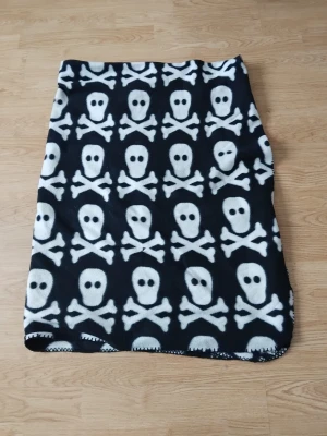  dödskallemönster filt  - Cool svart filt i fleece med vita dödskallar och korslagda ben över hela tyget. Mjuk och varm, perfekt för dig som gillar en edgy stil. Rak modell och kort längd, superenkel att matcha med en hoodie eller t-shirt.