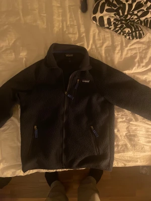 Mörkblå fleecejacka från Patagonia S - Mysig mörkblå fleecejacka från Patagonia i storlek S. Jackan har hel dragkedja framtill, två sidofickor med blå detaljer och hög krage. Perfekt för kyliga dagar och enkel att slänga på sig när du vill vara både bekväm och snygg.