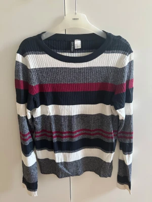 Randig ribbad tröja från H&M - Säljer en snygg långärmad tröja från H&M med breda ränder i vitt, grått, mörkblått och rött. Tröjan är ribbad och har en rund halsringning. Perfekt för dig som gillar färg och mönster i din garderob