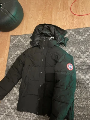 Svart dunjacka från Canada Goose - Säljer en svart dunjacka från Canada Goose med klassisk patch på ärmen. Jackan har huva, knäppning med både dragkedja och knappar samt quiltad design. Perfekt för kalla dagar och riktigt snygg till streetwear-stilen.