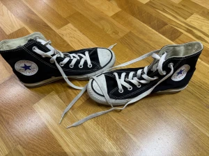 Svarta Converse Chuck Taylor High - Klassiska svarta Converse Chuck Taylor All Star high-top sneakers med vit sula och vita snören. Skorna har rund tå, platt sula och tydliga loggor på sidan och hälen. Tillverkade i slitstark bomull med kontrasterande vita sömmar och metallöglor för snörningen.