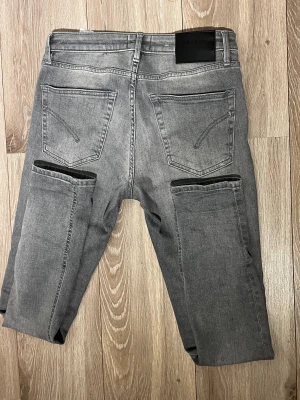 Grå jeans från Dondup, storlek 29 - Snygga grå jeans från Dondup, tillverkade i Italien. Modellen har raka ben och klassiska fem fickor, med coola detaljer på bakfickorna. Jeansen är i ett mjukt denimtyg och har en stilren look med svart patch bak i midjan.