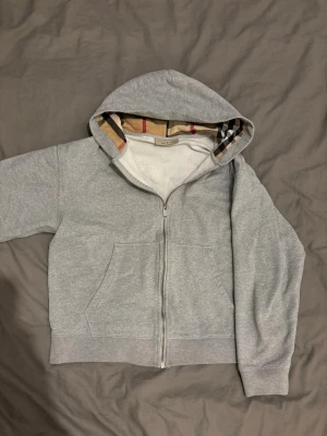 Grå Burberry hoodie med dragkedja - Snygg grå hoodie från Burberry i storlek M. Klassisk dragkedja framtill och två fickor. Huva med ikoniskt Burberry-rutigt foder i beige, svart och rött. Mjuk bomullskvalitet och broderad logga på bröstet. Perfekt för dig som gillar stilrena märkesplagg.