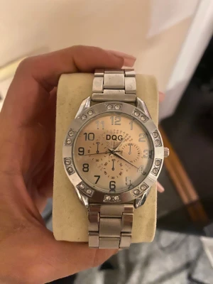 DQG silverfärgad klocka med stenar - Stilren klocka från DQG i silverfärg med metallarmband och rund urtavla. Urtavlan har tydliga siffror och dekorativa små stenar runt boetten som ger extra bling. Klockan har flera små urtavlor och en klassisk, tidlös design.