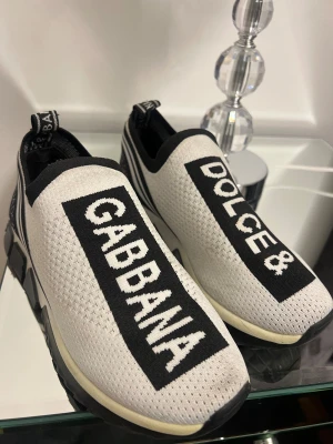 Dolce & Gabbana  sneakers Unisex - Snygga vita slip-on sneakers från Dolce & Gabbana med svart kontrast och tydlig logotyp på ovansidan. Skorna har en stickad ovandel i syntetmaterial, platt sula och svarta detaljer längs kanterna och hälen. Perfekt för dig som gillar statement-plagg och sportig stil.