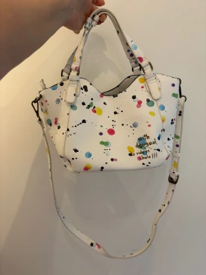 Vit handväska från Desigual med färgglada prickar - Säljer en vit handväska från Desigual med färgglada prickar i blått, rosa, gult och grönt. Väskan har både handtag och justerbar axelrem, samt cool textdetalj på framsidan. Perfekt för dig som gillar unika och lekfulla accessoarer.