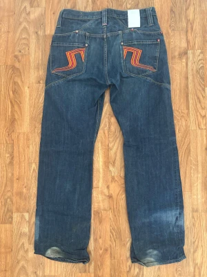 J.Lindeberg blå jeans med broderi - Snygga blå jeans från J.Lindeberg med coolt orange och rött broderi på bakfickorna. Klassisk femficksmodell med raka ben och kontrastsömmar. Röd detalj vid knapphål och nitar ger extra edge. Perfekt för dig som gillar street och sportig stil.