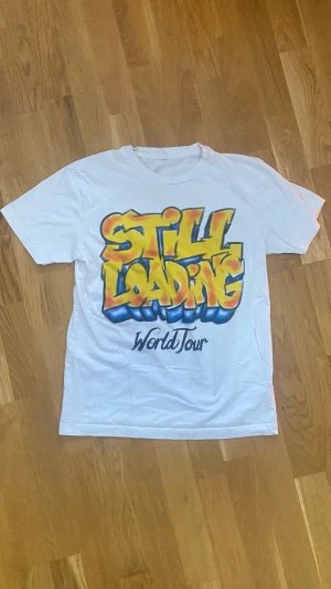 Vit t-shirt från Central Cees tour - Vit t-shirt från Central Cees koncert i Sverige. På framsidan står det 'Still Loading World Tour' i gult, orange och blått, och på baksidan finns en filmklappa med texten 'Live Yours'. Klassisk passform och rund halsringning.