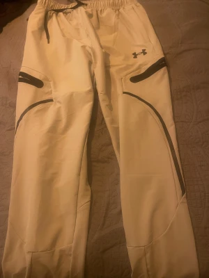Beiga täckbyxor från Under Armour - Säljer ett par beiga täckbyxor från Under Armour med svarta detaljer och snörning i midjan. Byxorna har elastisk midja och flera praktiska fickor med dragkedja. Perfekta för kalla dagar och outdoor-aktiviteter.