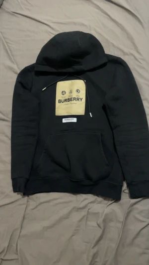 Svart Burberry hoodie med beige patch - Svart hoodie från Burberry med stor beige patch och logga på bröstet. Tröjan har huva med snörning, magficka och ribbade muddar. Perfekt för dig som gillar streetwear och vill sticka ut med ett lyxigt märke.