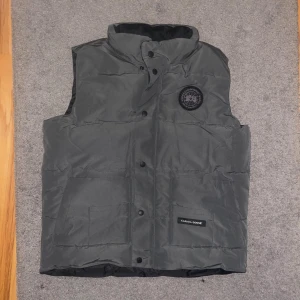 Mörkgrå dunväst från Canada Goose - Snygg mörkgrå dunväst från Canada Goose med hög krage och tryckknappar framtill. Västen har två stora fickor och ett svart Canada Goose-märke på bröstet. Perfekt för kalla dagar och har en stilren, clean look.