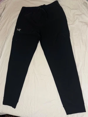 Svarta mjukisbyxor från Arc'teryx - Svarta mjukisbyxor från Arc'teryx med broderad logga på vänster lår. Byxorna har snörning i midjan, dragkedjeförsedda fickor och ribbade muddar vid bensluten. Sköna och stilrena, perfekta för chill eller träning.