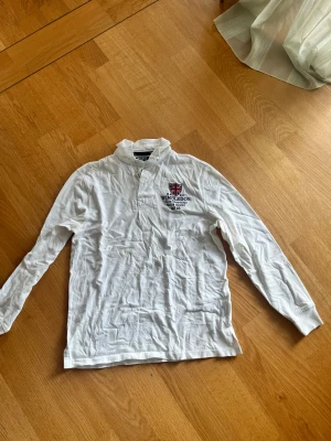 Vit Wimbledon henley från Polo Ralph Lauren - Vit långärmad henley från Polo Ralph Lauren med broderad Wimbledon-logga och brittisk flagga på bröstet. Skjortan är i 100% bomull och har knappar vid halsen. Snygg och sportig vibe, perfekt för dig som gillar klassisk stil med en touch av tennis. Funkar också får storlek S.