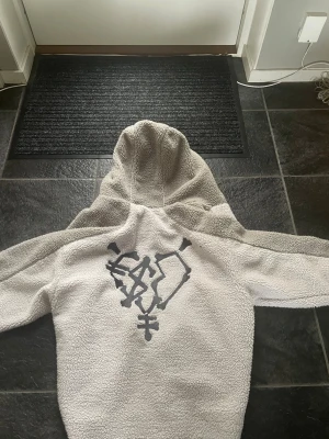 Teddyjacka från MoreMoneyMoreLove S - Säljer en fluffig teddyjacka från MoreMoneyMoreLove i storlek S. Jackan är vit och ljusgrå med huva, stora fickor framtill och broderad logga på bröstet. Baksidan har en unik svart grafisk design. Perfekt för dig som vill sticka ut med streetwear-vibe. As skön nu till vintern allt är helt nytt förutom lappen som man hänger upp den på är lite trasig men inget man märker, ny pris 119 euro vilket är 1368kr