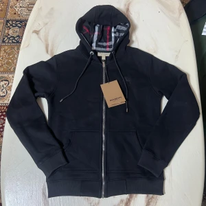Svart Burberry hoodie med dragkedja - Säljer en helt ny tjocktröja ifrån burberry. Den är svart och bockstavligen helt ny. Köpte den och den var alldeles för liten. Storleken är S och jag själv är 174cm lång samt 74kg och den var för liten. Inget fel med den annars.
