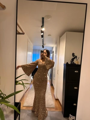 Glittrig guldig långklänning med paljetter - Säljer en maxad långklänning i guld med glittrande paljetter över hela klänningen. Den har vida trumpetärmar, figurnära passform och rundad ringning. Perfekt för dig som vill sticka ut och glänsa på bal eller fest. Materialet är syntet med massor av paljetter för extra wow-effekt.