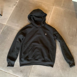 Svart hoodie från Polo Ralph Lauren - Svart hoodie från Polo Ralph Lauren med klassisk vit logga på bröstet. Tröjan har huva, magficka och ribbade muddar vid ärmslut och nederkant. Perfekt för chill dagar och enkel att matcha med jeans eller shorts. Mjuk och skön bomullskvalitet. Modellen är 180cm och väger 73kg