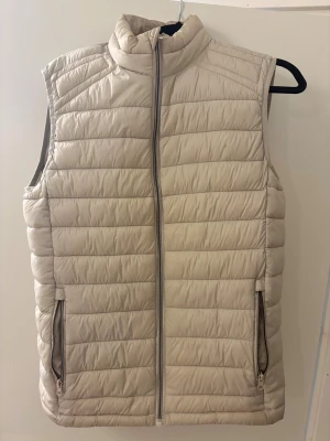 Beige dunväst från Blue Coast Yachting - Snygg beige dunväst från Blue Coast Yachting med quiltad design och hög krage. Västen har två praktiska fickor med dragkedja och en stilren, rak passform. Perfekt för lager-på-lager och enkel att matcha med olika outfits.