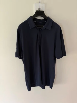 Mörkblå golfpiké från Galvin Green - Snygg mörkblå golfpiké från Galvin Green med klassisk krage och knappar framtill. Tillverkad i mjuk bomull och har korta ärmar. Diskret logga på ärmen och i nacken. Perfekt för en clean och sportig stil. Storlek: L passar M. Nypris: 999kr