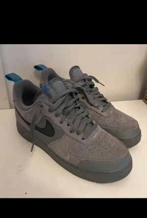 Nike Air Force 1 grey (winter edition) - Nike Air Force 1 sneakers i grå mocka med mörkgrå detaljer och svart swoosh. Låg modell med snörning och platt sula. Blå Nike-logga på tungan och blå flikar bak. Perfekt för dig som gillar stilrena och klassiska sneakers med en twist.