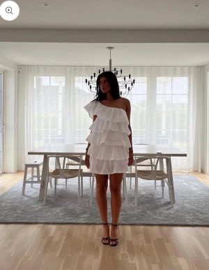 Studentklänning med volanger - Säljer en vit kortklänning med flera lager volanger och oneshoulder-design. Klänningen har en omlott-look och är axelbandslös på ena sidan, vilket ger en unik och trendig siluett. Perfekt för dig som gillar statement-plagg med mycket rörelse och volym.