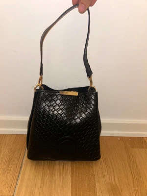 Svart axelväska i skinn med guldiga detaljer - Snygg svart axelväska i skinn med flätad struktur och guldiga metalldetaljer. Väskan har en stilren form och ett kort axelband som ger en trendig look. Perfekt för dig som vill ha en elegant och tidlös accessoar.