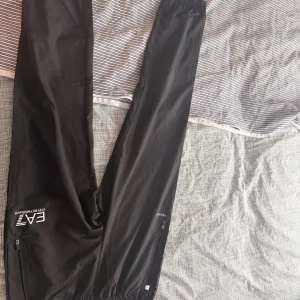 Svarta täckbyxor EA7 Emporio Armani - Svarta täckbyxor från EA7 Emporio Armani med elastisk midja och dragkedjor vid fickorna. Byxorna har en stilren look med logga på benet och är gjorda i lätt syntetmaterial som passar perfekt för utomhusaktiviteter. Snyggt sportig vibe.