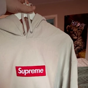 Beige Supreme hoodie med box logo - Supreme hoodie i beige med klassisk röd box logo framtill. Tröjan har huva med snörning. Perfekt för dig som gillar streetwear och vill sticka ut med en ikonisk look.