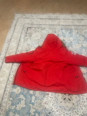 Röd North Bend parkas med fuskpäls - Säljer en röd parkas från North Bend med huva och fuskpälsdetalj. Jackan har flera fickor, dragkedja och Aqua-Tex-tyg som står emot vatten. Insidan är svart och vadderad för extra värme. Perfekt för kalla dagar och har en schysst streetstil.