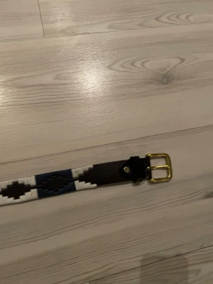 Pampeano bälte - Säljer ett mörkbrunt skärp i skinn med coolt geometriskt mönster i vitt och marinblått. Skärpet har en guldfärgad metallspänne och känns riktigt stilrent till jeans eller chinos.