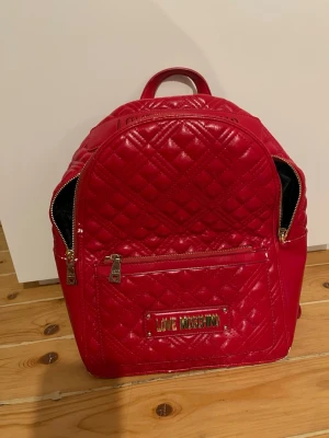 Röd quiltad ryggsäck Love Moschino -  Röd ryggsäck från Love Moschino. Väskan är quiltad med guldfärgade dragkedjor och detaljer, har ett stort huvudfack samt en praktisk ytterficka. Materialet är glansigt syntet och insidan har logomönstrat foder.