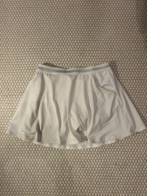 Vit kortkjol från Artengo - Säljer en vit kortkjol från Artengo med glittrigt silverband i midjan. Kjolen har en lätt utsvängd passform och är gjord i ett mjukt, stretchigt syntetmaterial som passar perfekt till tennis eller andra sporter.