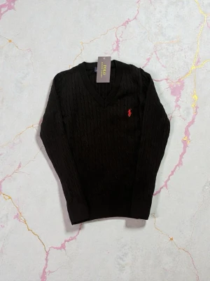 Svart kabelstickad tröja Polo Ralph Lauren - Kabelstickad svart tröja från Polo Ralph Lauren med klassisk v-ringning och röd broderad logga på bröstet. Långärmad modell i mjuk bomull som passar perfekt till både jeans och kjol. Snygg ribbad kant vid ärmslut och nederkant.