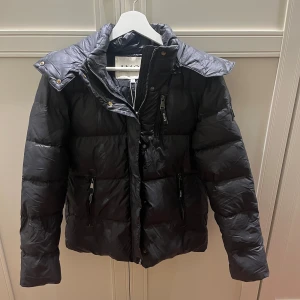 Svart pufferjacka, design by Amor  - Säljer en helt ny svart pufferjacka från design by Amor i storlek S. Jackan har hög krage, dragkedja framtill och flera fickor med dragkedjor. 