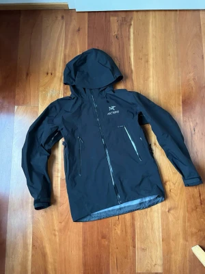 Svart vindjacka från Arc'teryx - Säljer en svart vindjacka från Arc'teryx med huva och flera dragkedjeförsedda fickor. Jackan har en clean look med logga på bröstet och justerbara muddar. Perfekt för dig som gillar outdoor och vill ha något funktionellt och stilrent. Jackan har ett litet hål bredvid vänster ficka och den andra linningen är trasig men den fungerar bra.